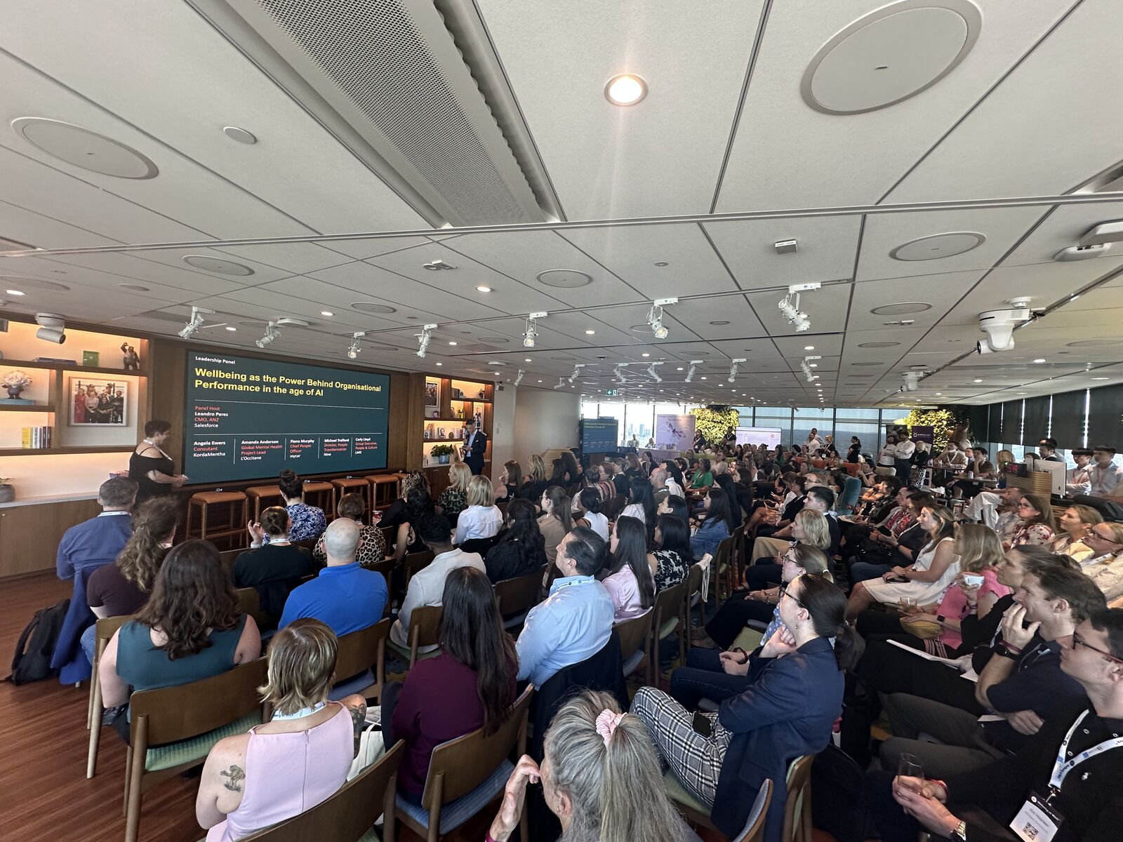 Packed conference room at the Wellbeing at Work Summit — slide on screen reads "Wellbeing as the Power Behind Organisational Performance in the Age of AI"