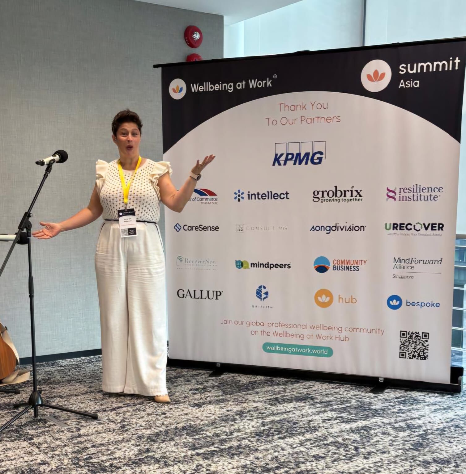 Amanda Anderson at the Wellbeing at Work Summit Asia, arms raised, standing in front of the KPMG and partner sponsor board