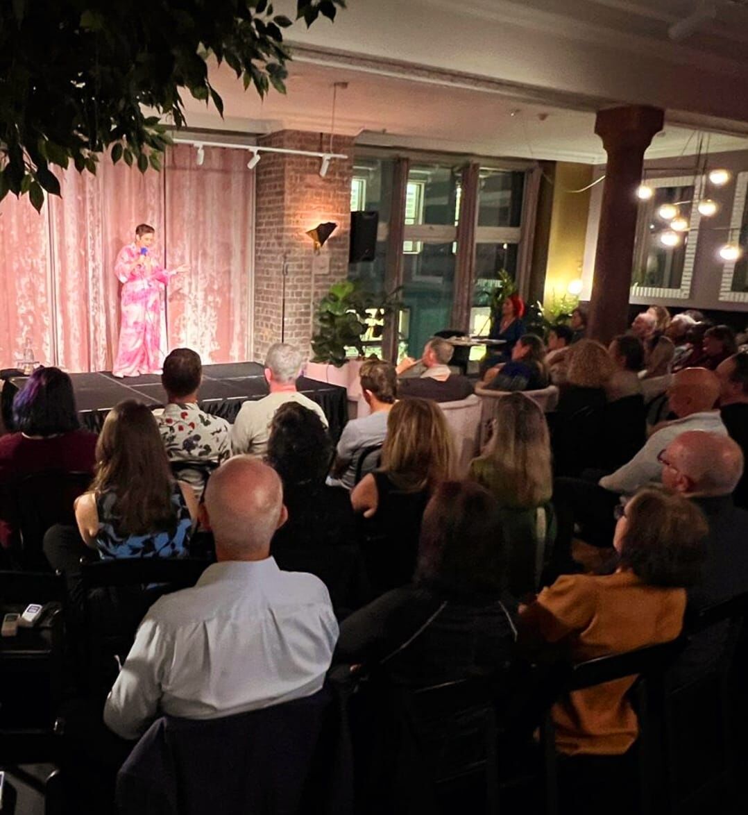Amanda Anderson on a warmly lit stage in a pink outfit, speaking to a packed intimate venue