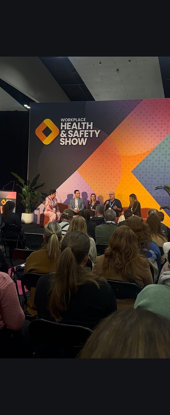 Five-person panel discussion on stage at the Workplace Health & Safety Show, with Amanda Anderson far left, full audience in foreground